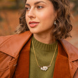 A woman with short, wavy brown hair wears a brown leather jacket over a green sweater and an elegant Tulip Necklace with a white floral pendant, standing outdoors with an autumn background.