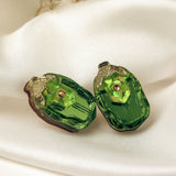 Two green, oval-shaped glass craft beads with a faceted surface and a small pink center gem, perfect for nature-inspired earrings, resting on a white silky fabric backdrop with blurred dried flowers in the background.