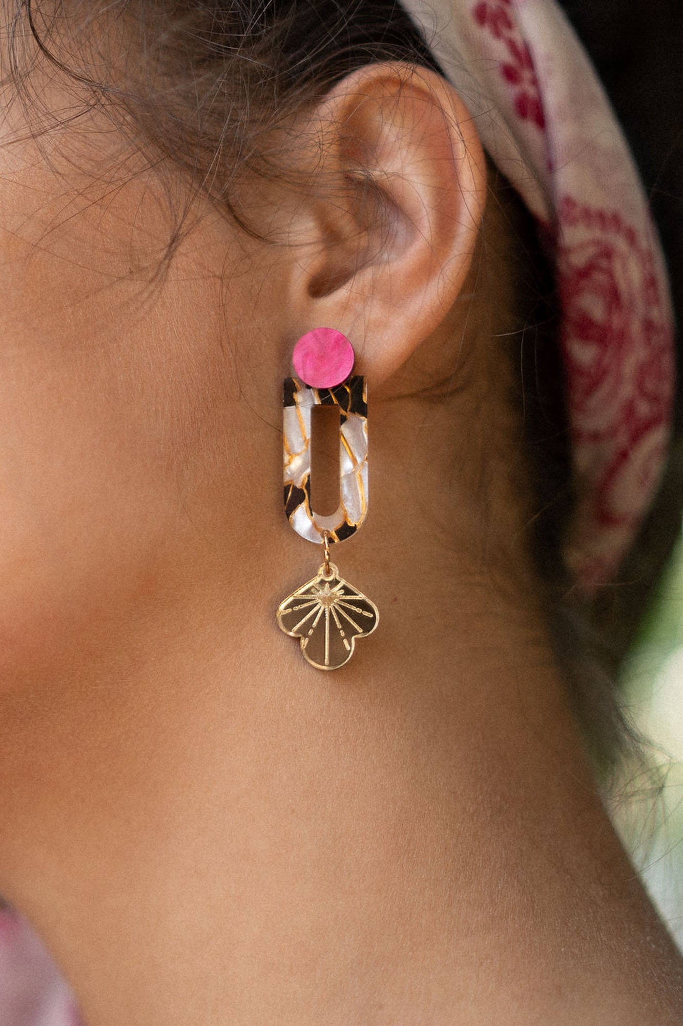 Close-up of a womans ear wearing Sakura Earrings—a statement earring with a bright pink stud, tortoiseshell oval link, and gold floral charm. A pink patterned headscarf adds an extra touch of elegance and artistry to the look.