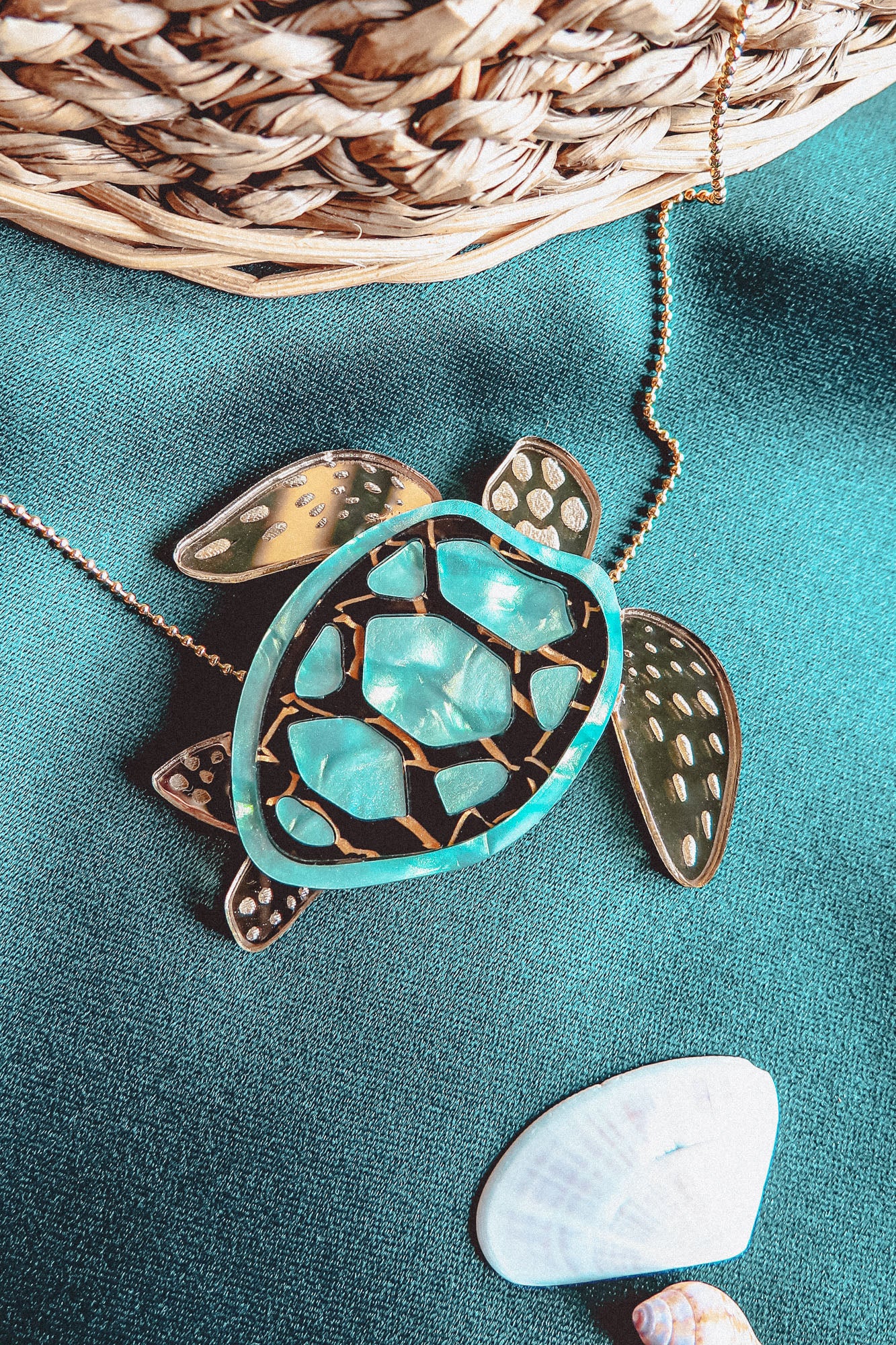 A close-up of a handcrafted Sea Turtle Necklace pendant with a turquoise and gold shell, lying on green fabric next to a white seashell and a wicker basket.
