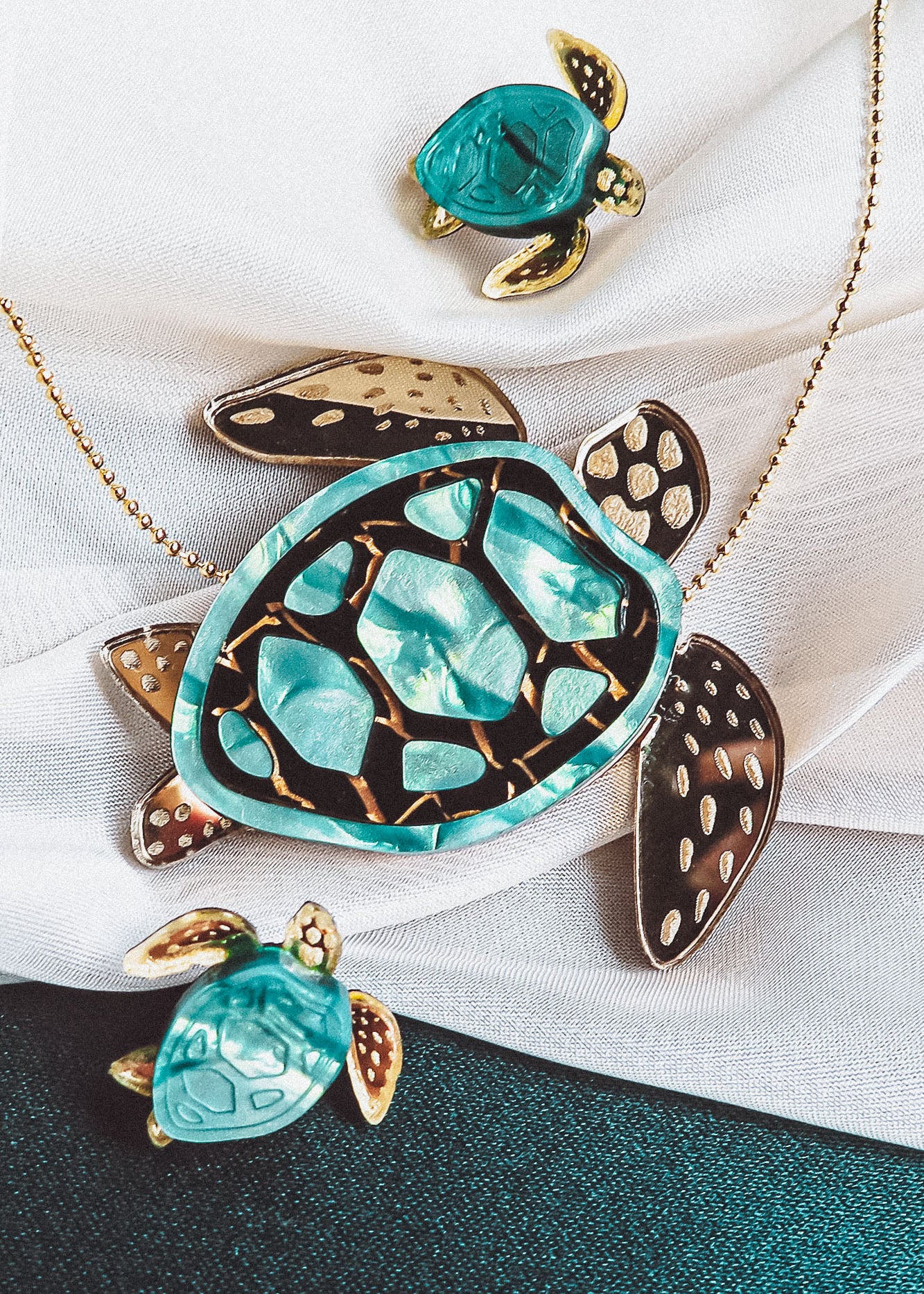 A handcrafted gold necklace featuring a turquoise and gold sea turtle pendant, accompanied by two matching small turtle charms, beautifully displayed on white fabric.