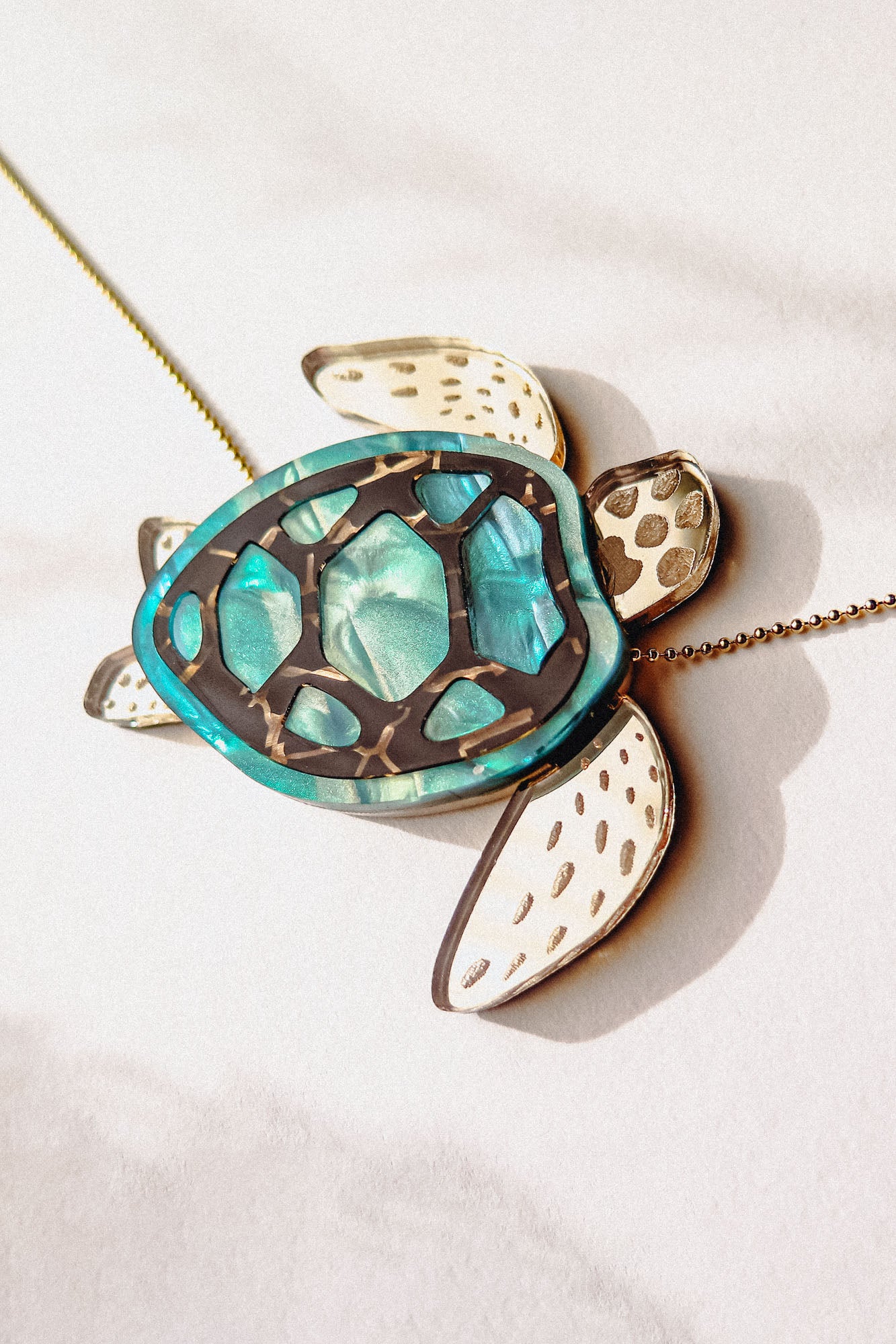 A close-up of a handcrafted Sea Turtle Necklace with a turquoise shell and white flippers, lying on a light surface. The turtle has intricate shell details and a gold chain.