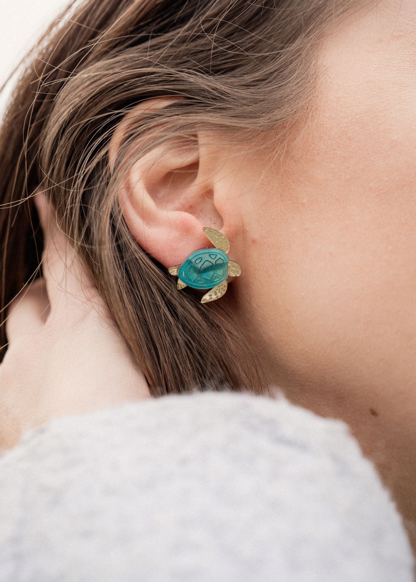 A close-up of a persons ear wearing gold and turquoise Sea Turtle Studs, with their light brown hair partially visible, captures an ocean-inspired, summery vibe.