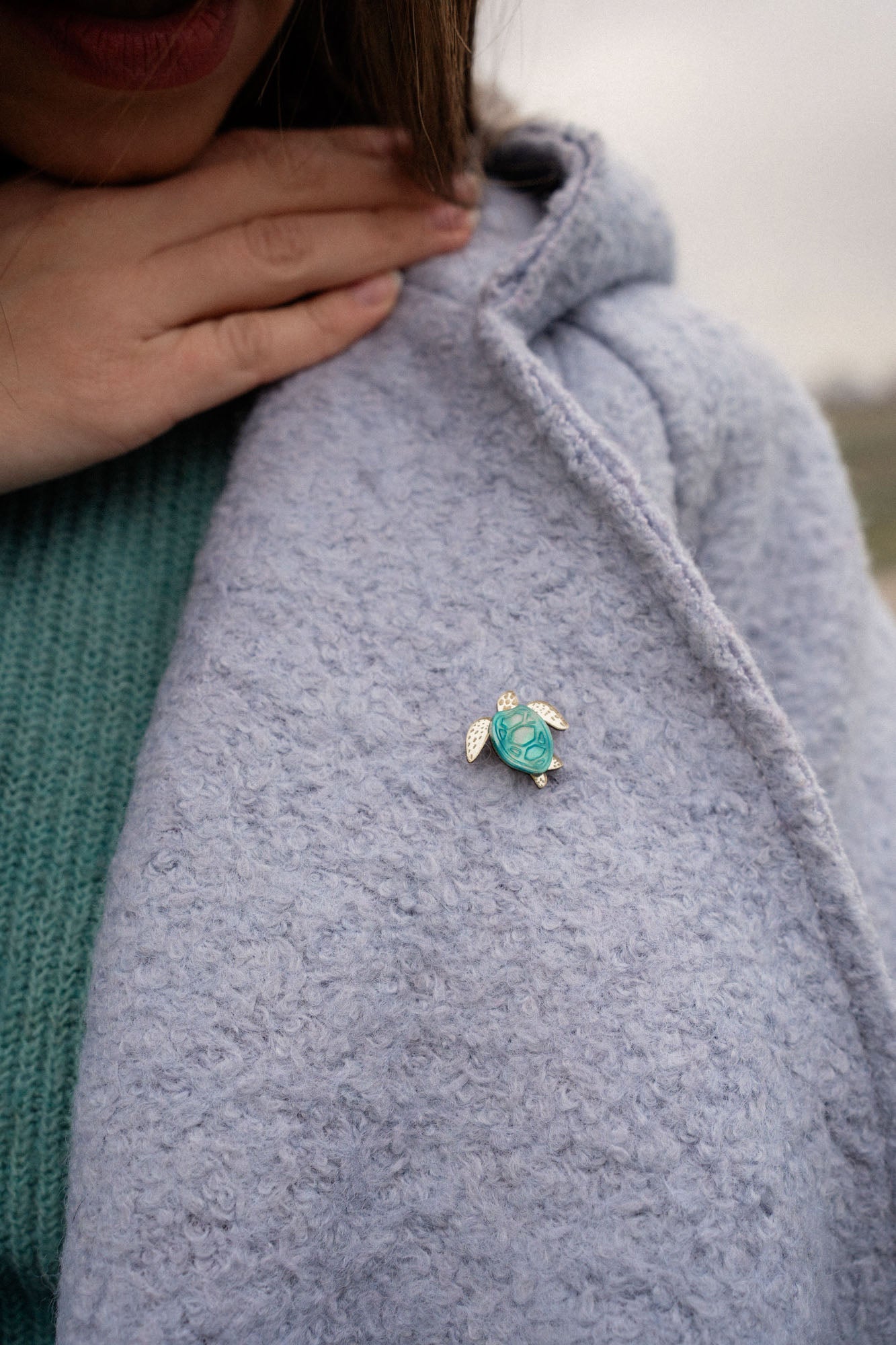 A person wearing a light blue textured coat with a handcrafted sea turtle pin in turquoise and silver on the lapel, and a green sweater underneath. Their hand is gently resting near the collar.