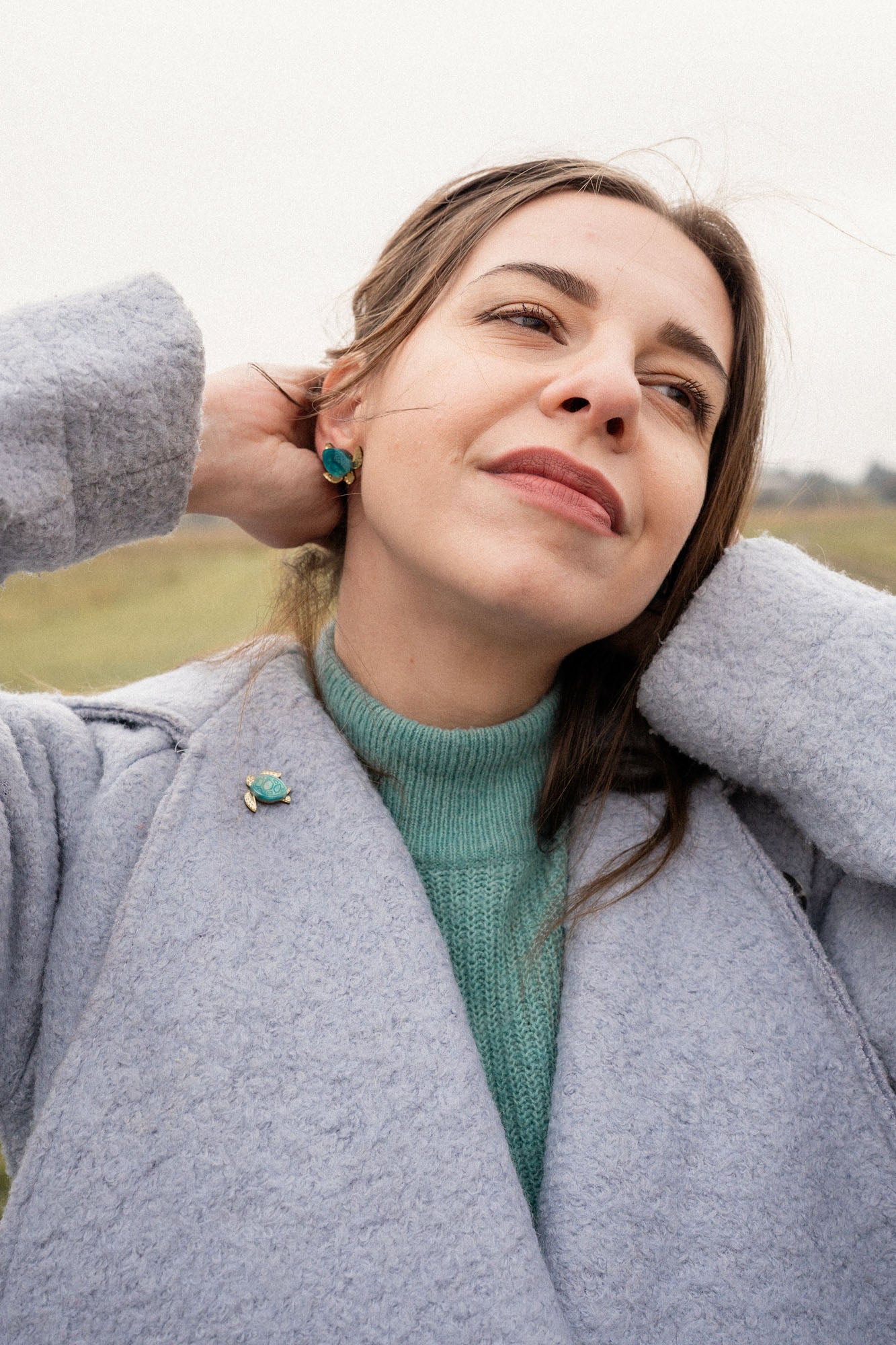 A woman in a light blue coat and teal sweater stands outdoors, smiling gently with her hands behind her head. She wears matching turquoise earrings and a handcrafted sea turtle pin made of mirror-acrylic glass, with a grassy field in the background.