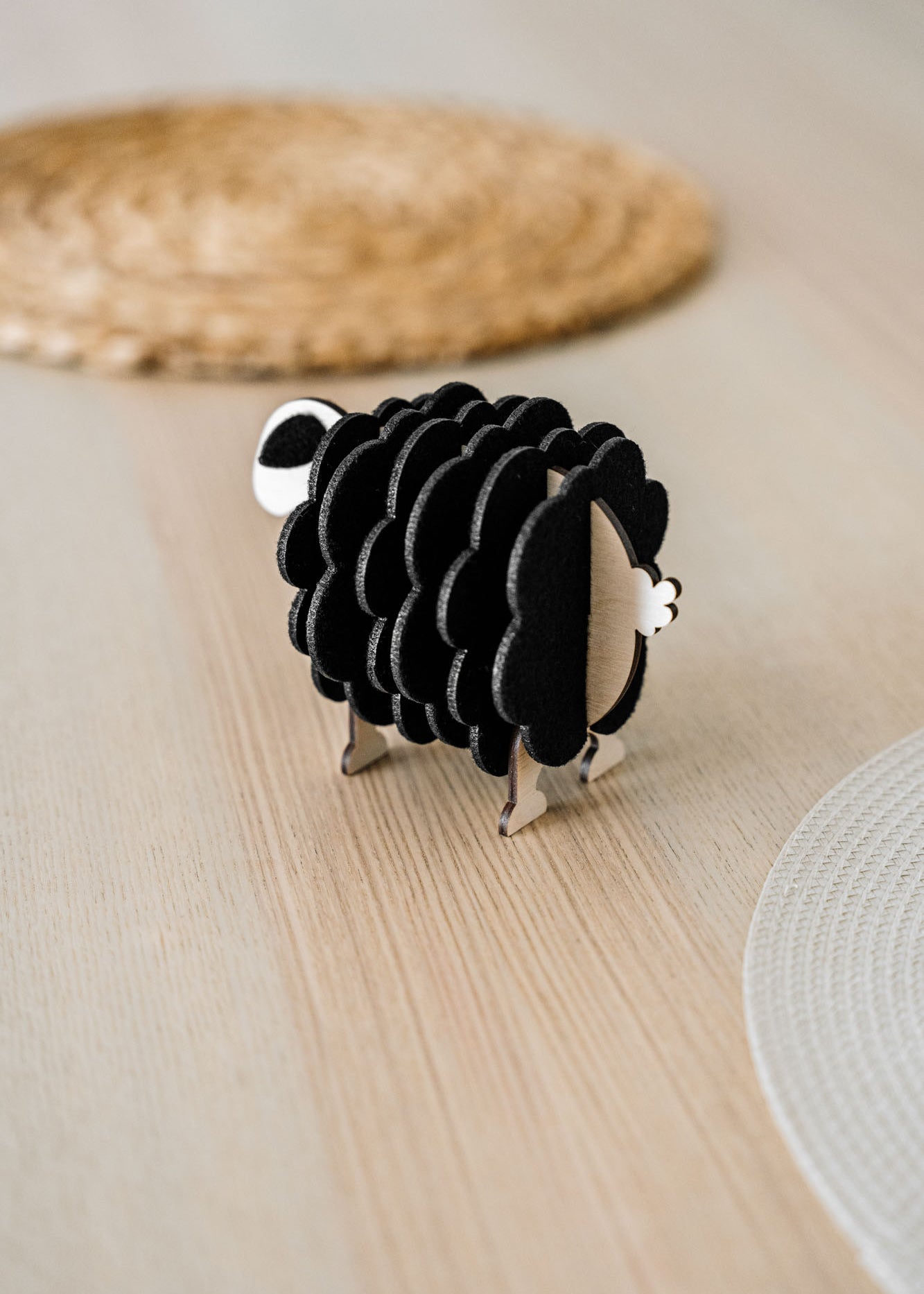 A small, black and white wooden sheep-shaped figure sits on a light wooden table near woven placemats, adding a charming touch to your home decor.