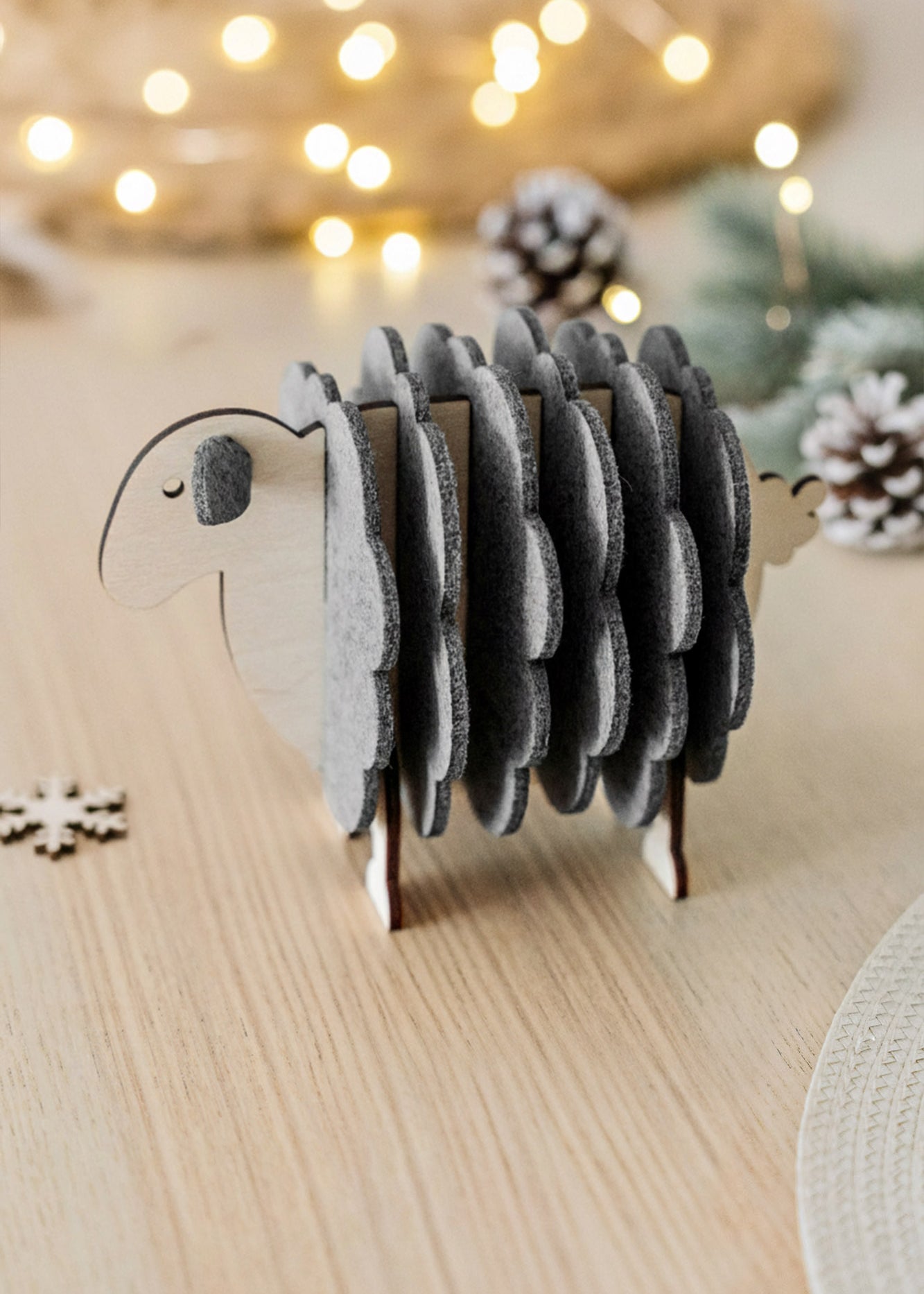 A wooden sheep-shaped coaster holder with felt coasters sits on a light wooden table, surrounded by pinecones, a snowflake decoration, and softly glowing fairy lights—charming home decor for cozy gatherings.