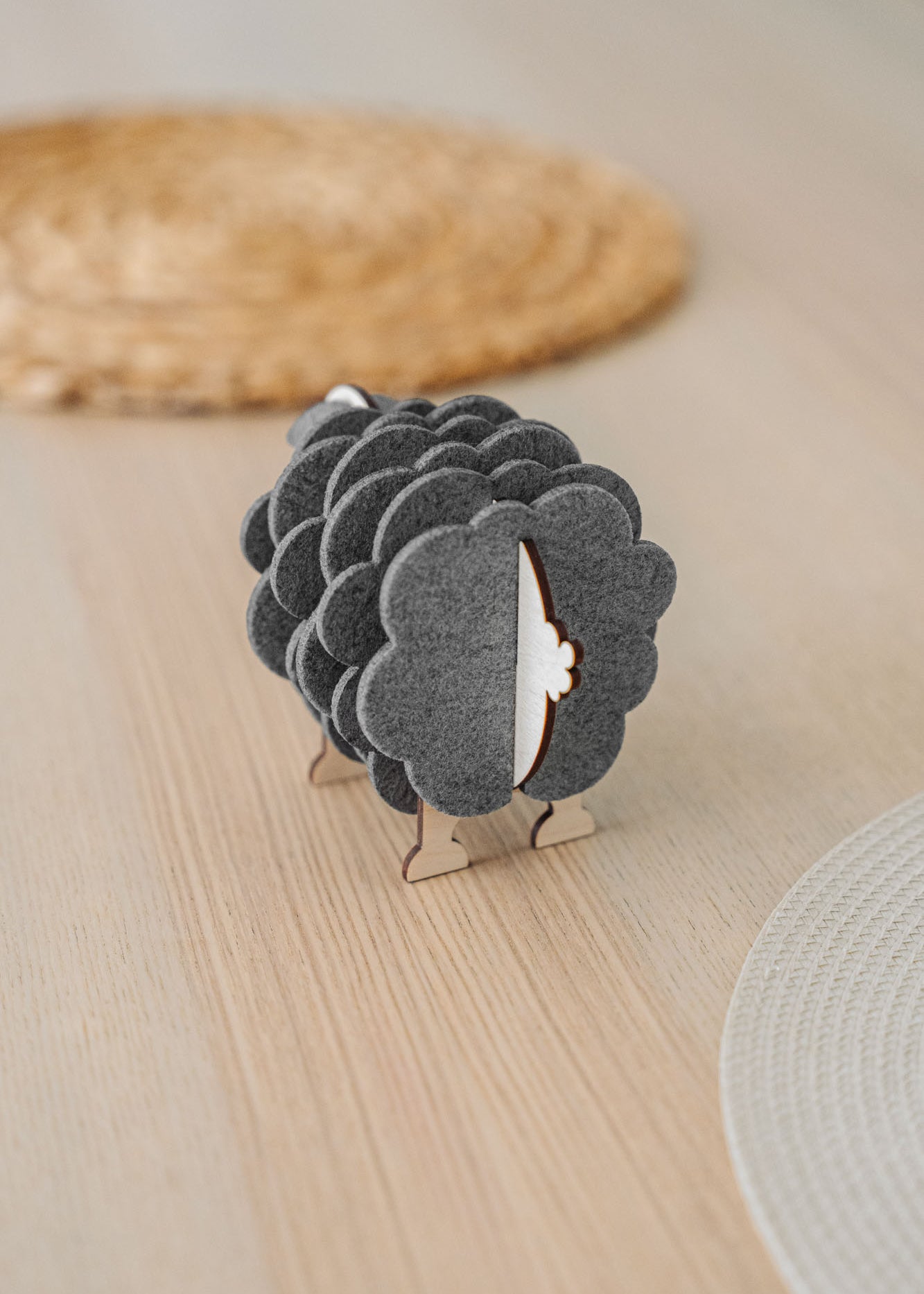 A wooden sheep-shaped coaster holder with dark felt coasters stands on a light table, adding a charming touch to your home decor. Woven placemats are visible in the background.