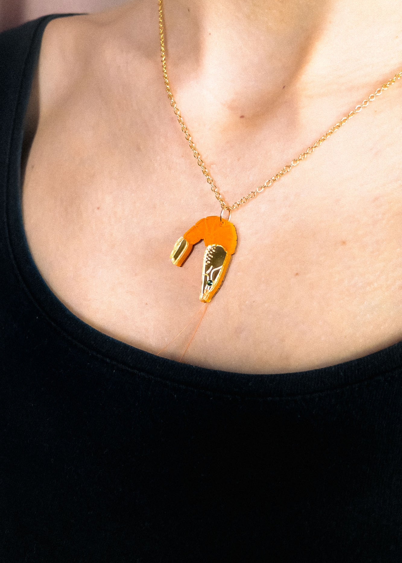 A whimsical Shrimp Pendant Necklace with a gold chain features an orange shrimp charm, playfully displayed above a black top on the wearer’s neck.