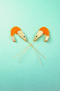 Two neon orange Shrimp Stud earrings, shaped like shrimp with long antennae crossing in the center, are positioned facing each other on a turquoise background. Subtle emerald rhinestones add a touch of sparkle to their playful design.