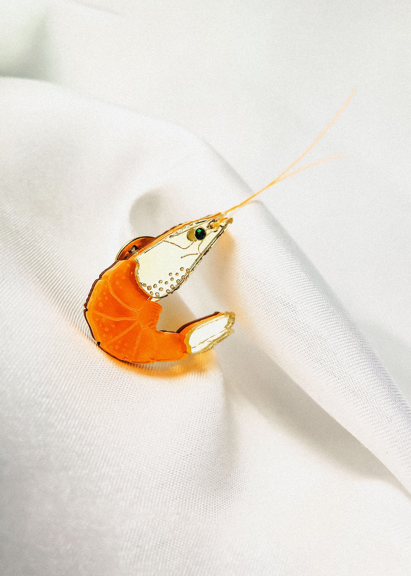 A decorative orange and clear glass shrimp figurine, perfect as an accessory or unique wardrobe charm, rests on white, softly folded fabric, showcasing fine details on its body and long antennae extending outward.