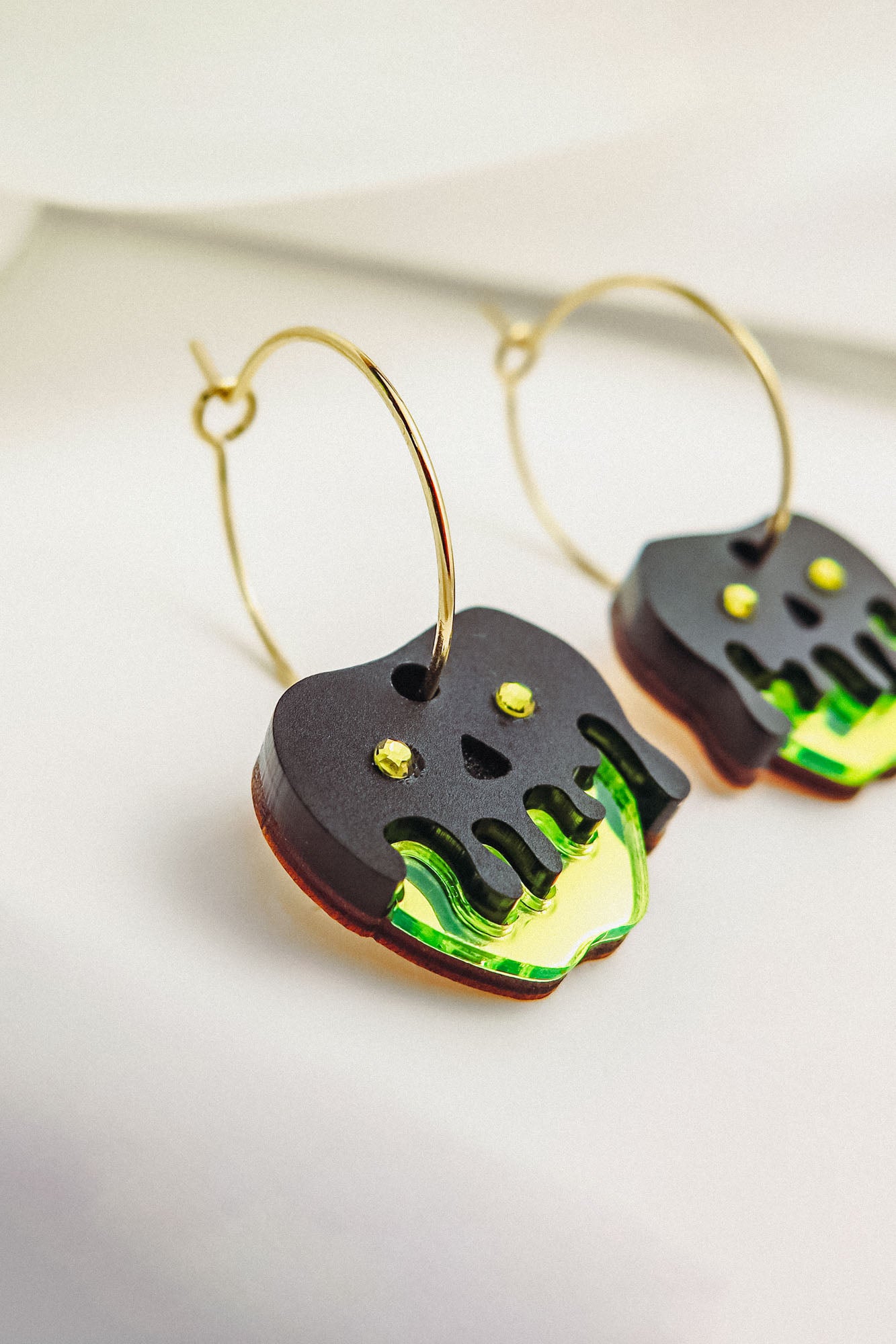 Limited edition Poisoned Apple Earrings with gold hoops and dangling black and neon green charms. Featuring a Halloween design with stylized dripping details and yellow dot accents, these earrings rest elegantly on a white surface.