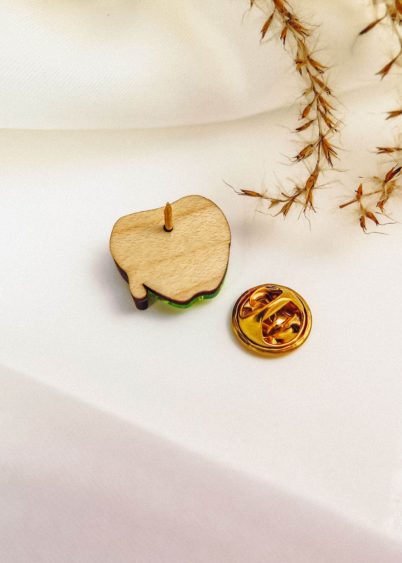 A wooden enamel Poisoned Apple Badge pin, shown from the back with its gold butterfly clutch beside it, rests on a white surface with dried grass in the corner—perfect for Halloween lovers. Limited stock available!.