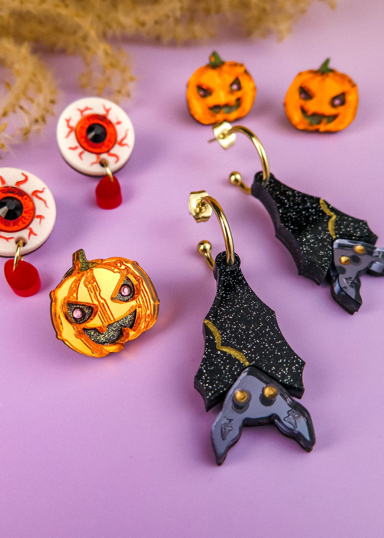 A set of Halloween-themed, hand-painted earrings on a purple surface, including pumpkin and eyeball earrings, a pumpkin brooch, and black bat-shaped earrings with glitter and purple accents. Dried plants are partially visible at the top.