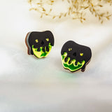 A pair of handcrafted earrings shaped like black skulls with glowing green drips at the bottom, perfect for a bold Halloween look, displayed on a white surface with dried grass in the background.