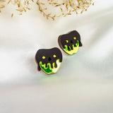 Two cookies shaped like apples with black and neon green dripping icing, resembling a poison apple, showcase a handcrafted Halloween look as they sit on a white fabric surface with delicate dried grass in the background.