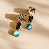 A pair of handcrafted geometric earrings with gold angular tops, black middle sections, and blue tear-drop pendants, displayed on a light beige surface with soft shadow patterns.