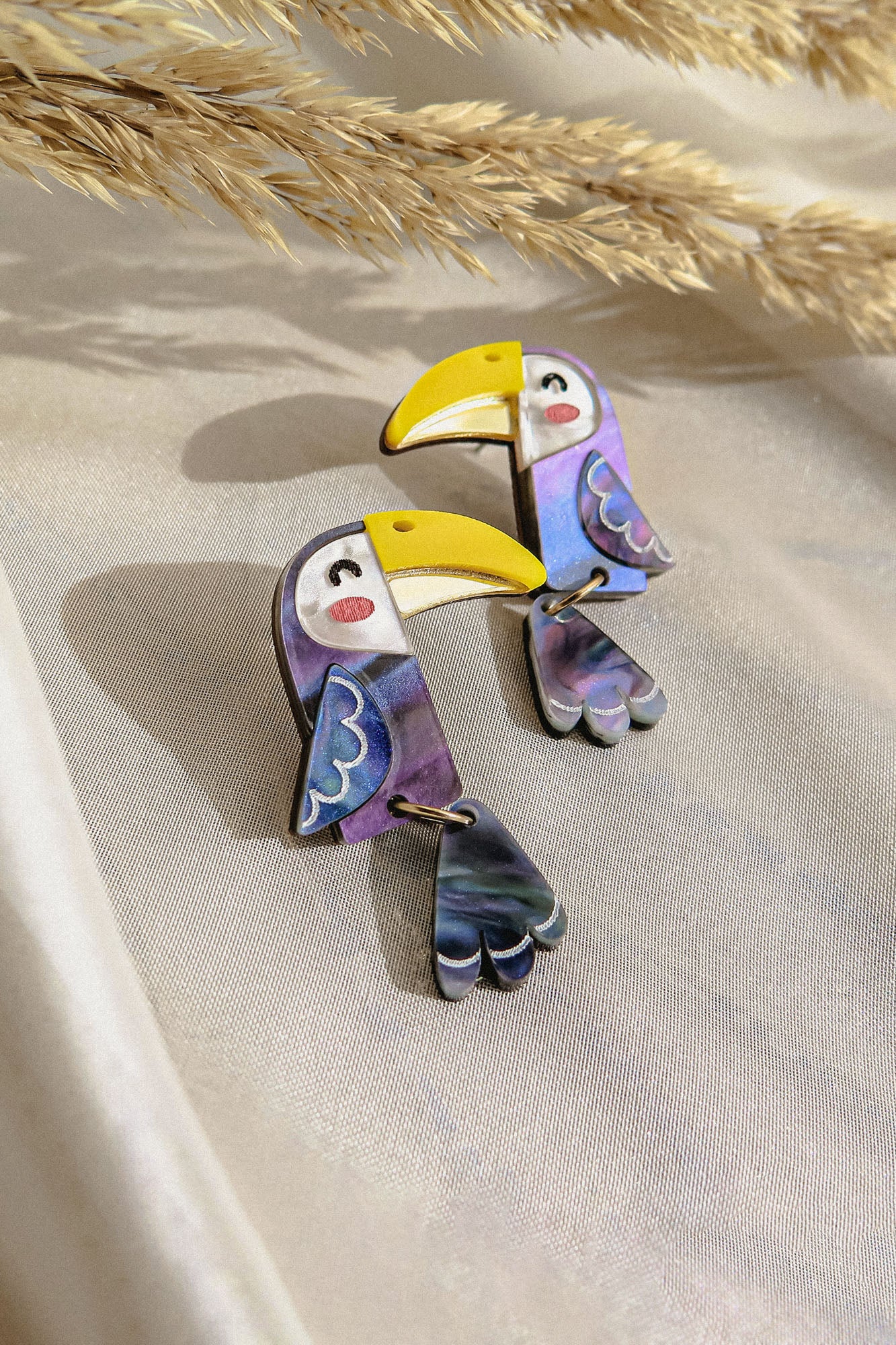 Handcrafted Toucan Earrings featuring yellow beaks, purple and blue wings, and dangling tails add a playful pop of color. Displayed on a light fabric background with dried pampas grass in the corner.