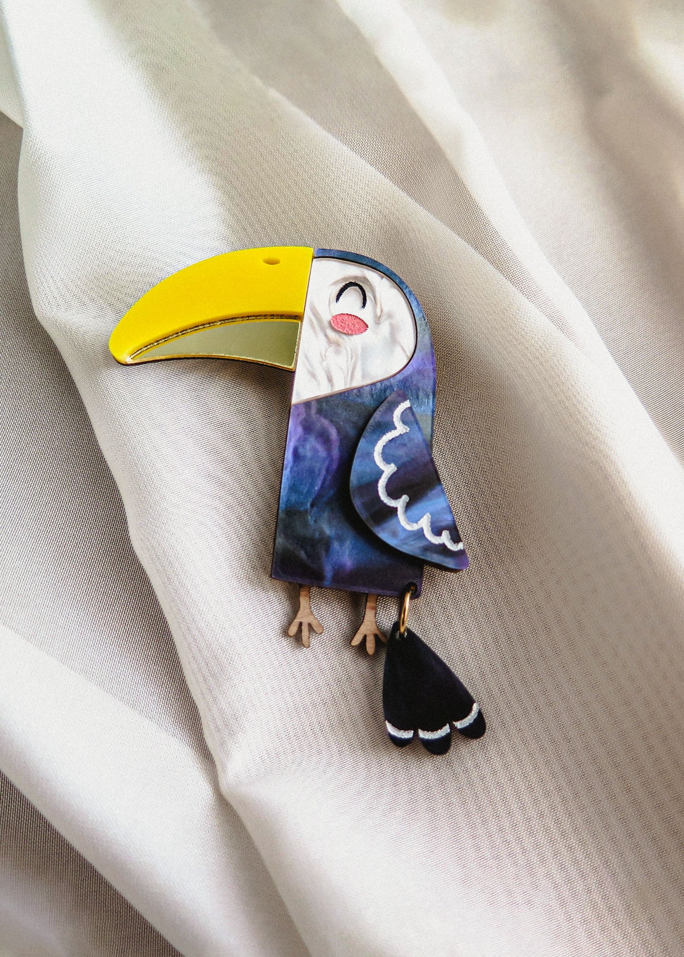 A handcrafted Toucan Brooch with a bright yellow beak and dark blue body captures tropical charm as it rests on soft, white, textured fabric.