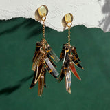 Featherlight gold and tortoiseshell dangle earrings with layered, leaf-like charms hang from a white textured surface above a green background. Light casts soft shadows on the wall.