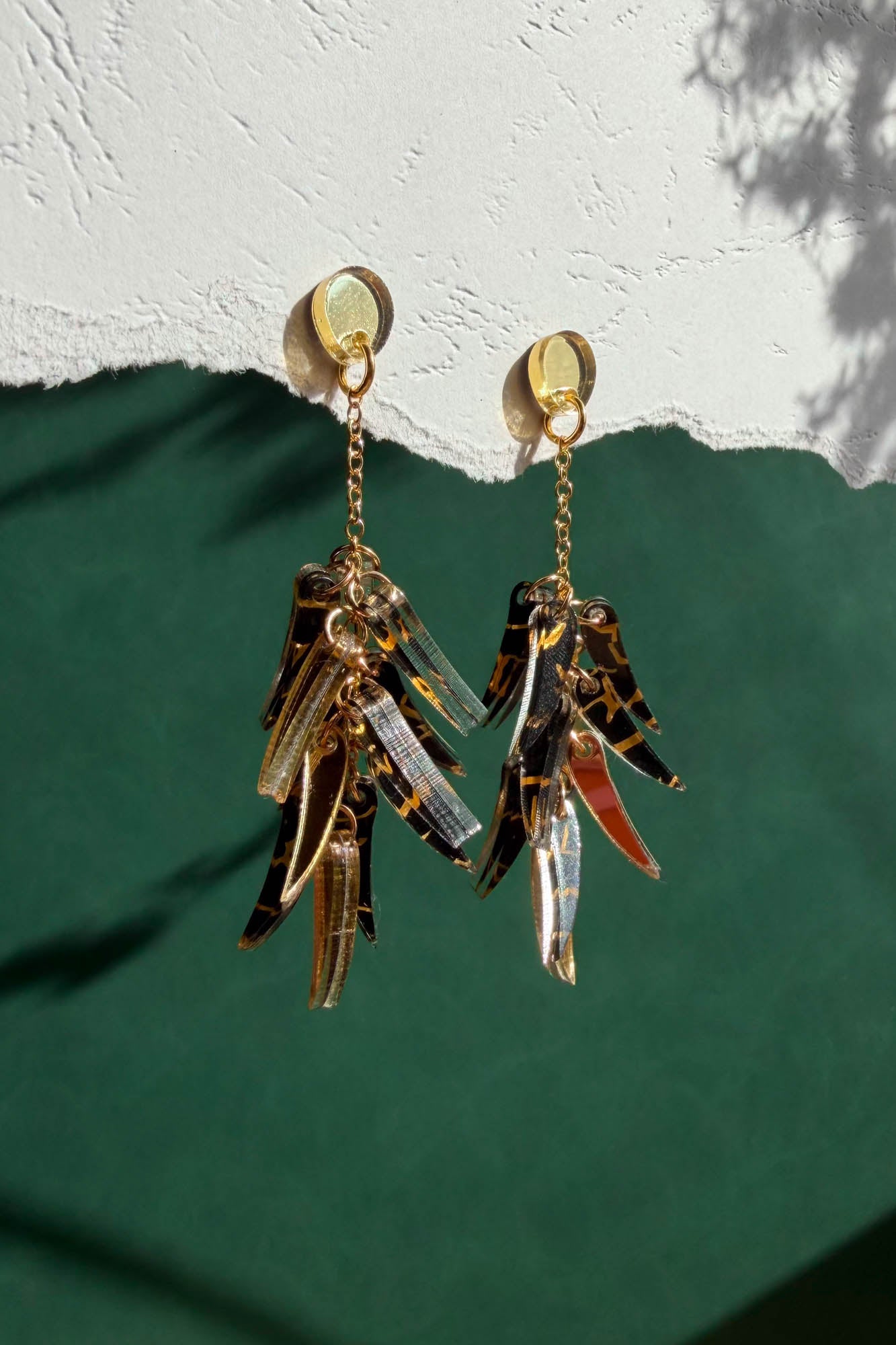 Featherlight gold and tortoiseshell dangle earrings with layered, leaf-like charms hang from a white textured surface above a green background. Light casts soft shadows on the wall.