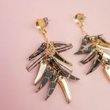 Gold and black marbled featherlight dangle earrings with multiple layered, leaf-shaped pendants arranged in a fan shape, displayed on a soft pink background.
