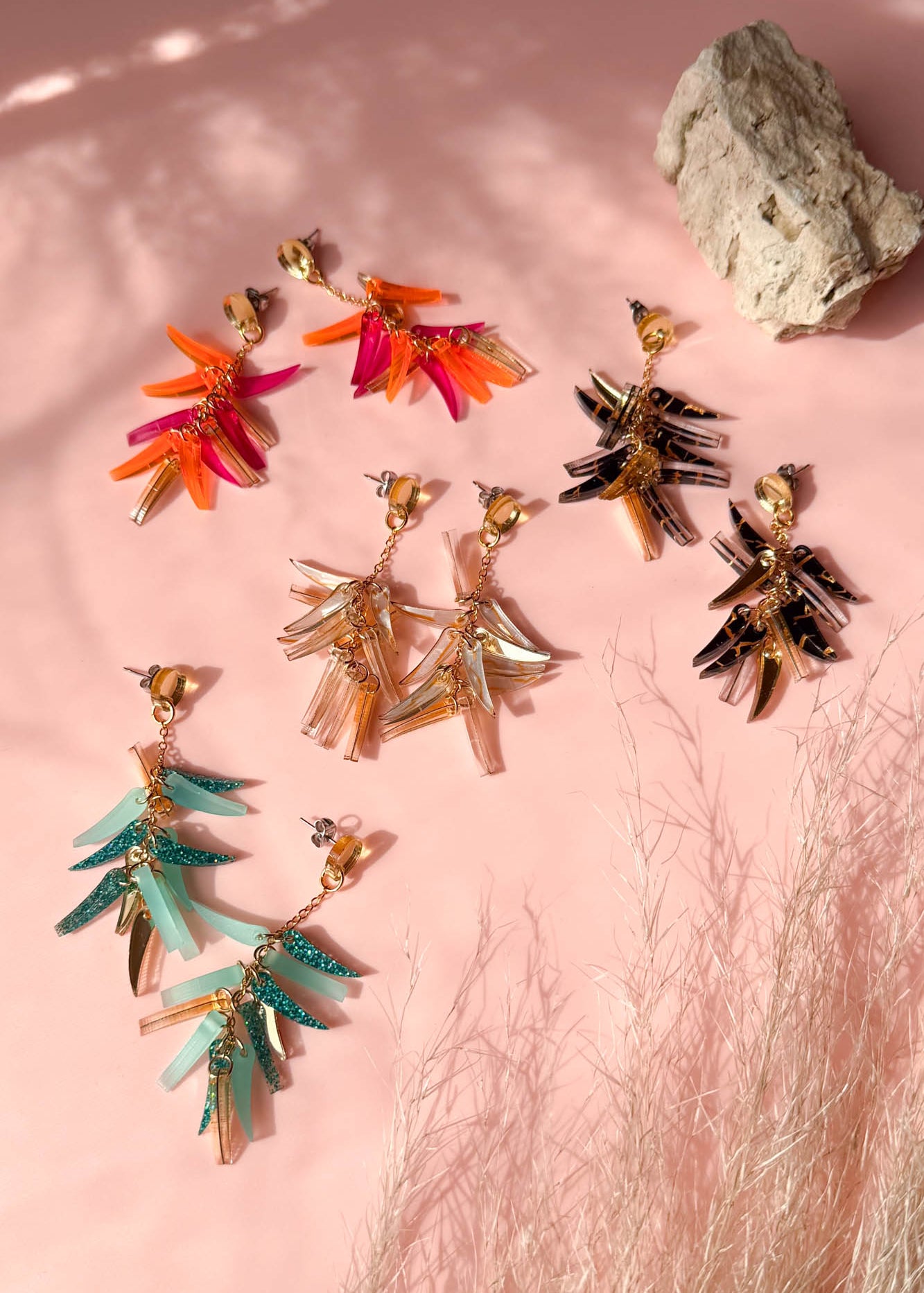 Colorful featherlight dangle earrings in four pairs—red-orange, brown-black, teal, and beige—feature acrylic tufts and are arranged on a pink surface with natural light, a beige rock, and wispy dried grass in the background.