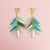 A pair of dangling earrings with gold hardware, featuring layered teal, blue glitter, and gold leaf-shaped charms, these unique style earrings are displayed on a light pink background.