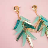 A pair of gold dangle earrings with teal and glittery blue acrylic accents, these unique style earrings are displayed on a pale pink background.
