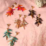 Five pairs of colorful, leaf-shaped acrylic earrings are arranged on a pink surface with shadows and a rock in the background. The earrings showcase a unique style in shades of red, orange, gold, black, and turquoise.