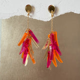 A pair of featherlight dangle earrings with gold studs and chains, each featuring clusters of translucent pink, orange, and clear acrylic shards, displayed against a textured white and metallic gold background.