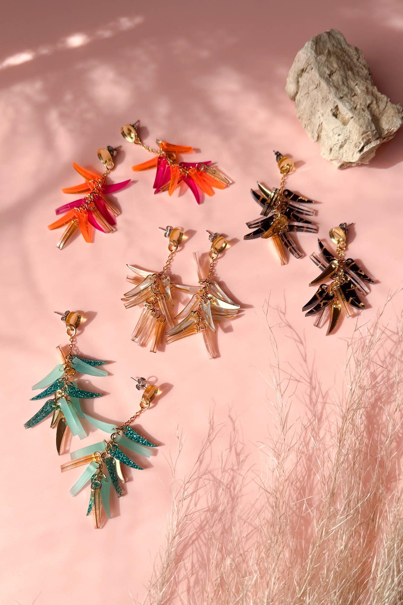 Colorful featherlight dangle earrings in four pairs—red, orange, teal, and black—are arranged on a pink surface with a rock and delicate dried grass, all showcased in natural sunlight.