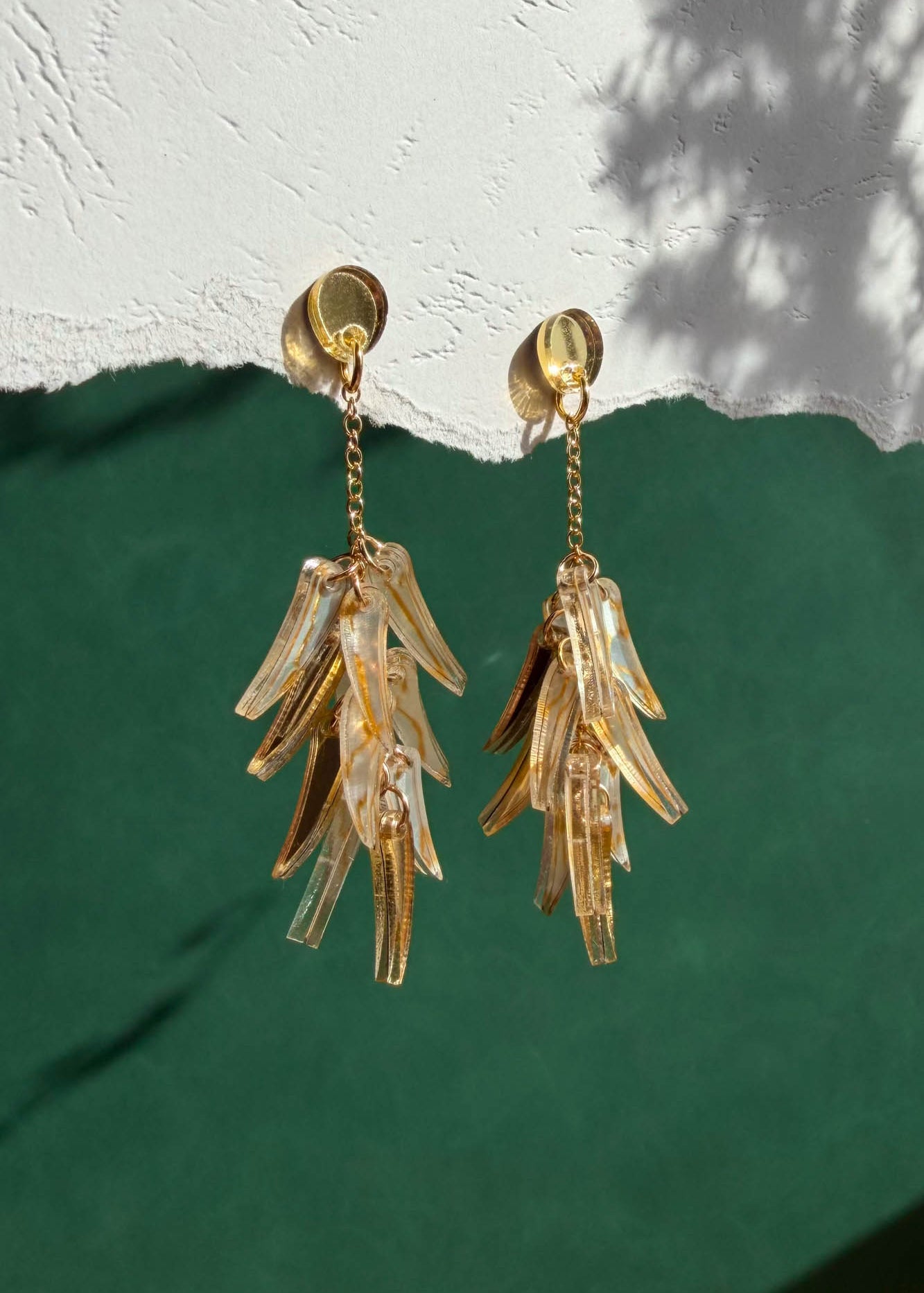 A pair of featherlight, gold chain dangle earrings with translucent, leaf-shaped pendants hang against a textured white and green background with shadow patterns.