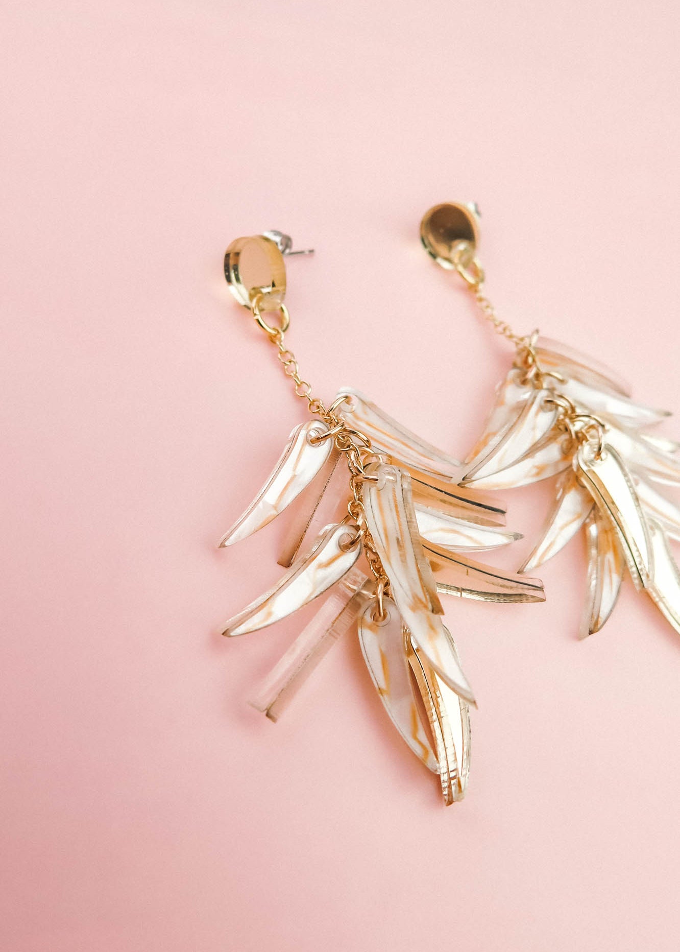 A pair of featherlight gold dangle earrings with leaf-shaped, translucent accents, displayed on a soft pink background.