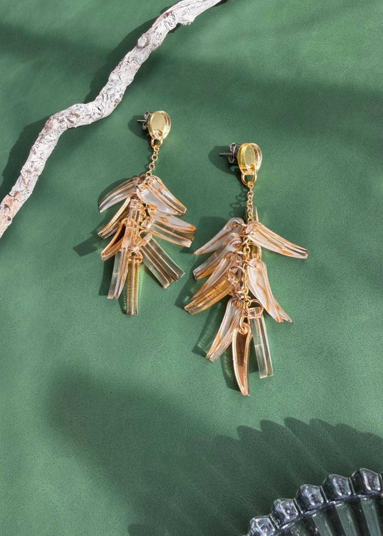 A pair of gold and clear acrylic dangle earrings shaped like layered leaves, these featherlight earrings are displayed on a green surface with textured driftwood and a partial glass dish.