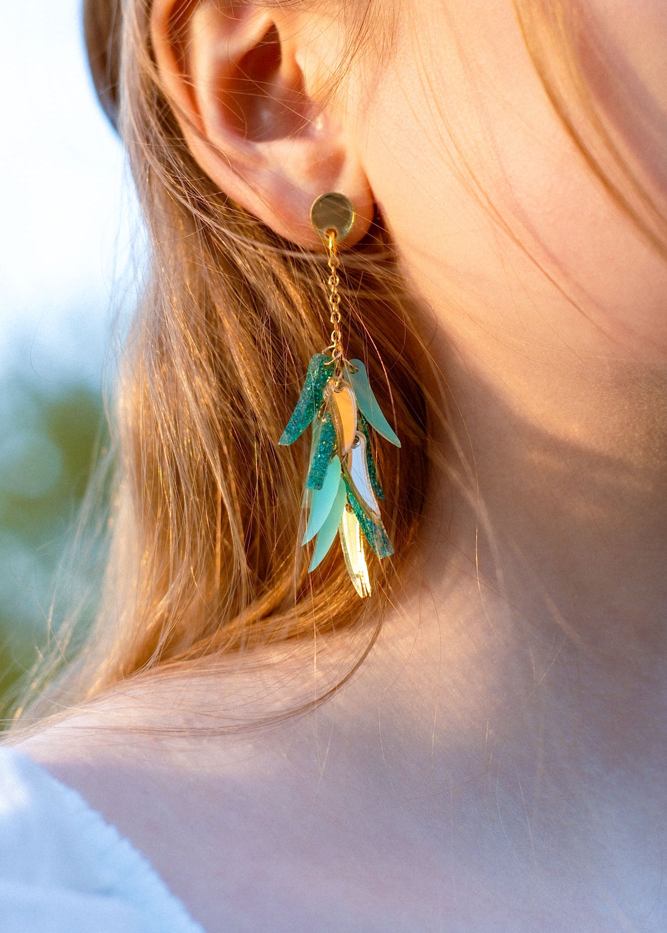 Close-up of a person wearing featherlight gold stud dangle earrings with metallic and turquoise leaf-shaped charms. The person has light brown hair, fair skin, and is dressed in a white top.