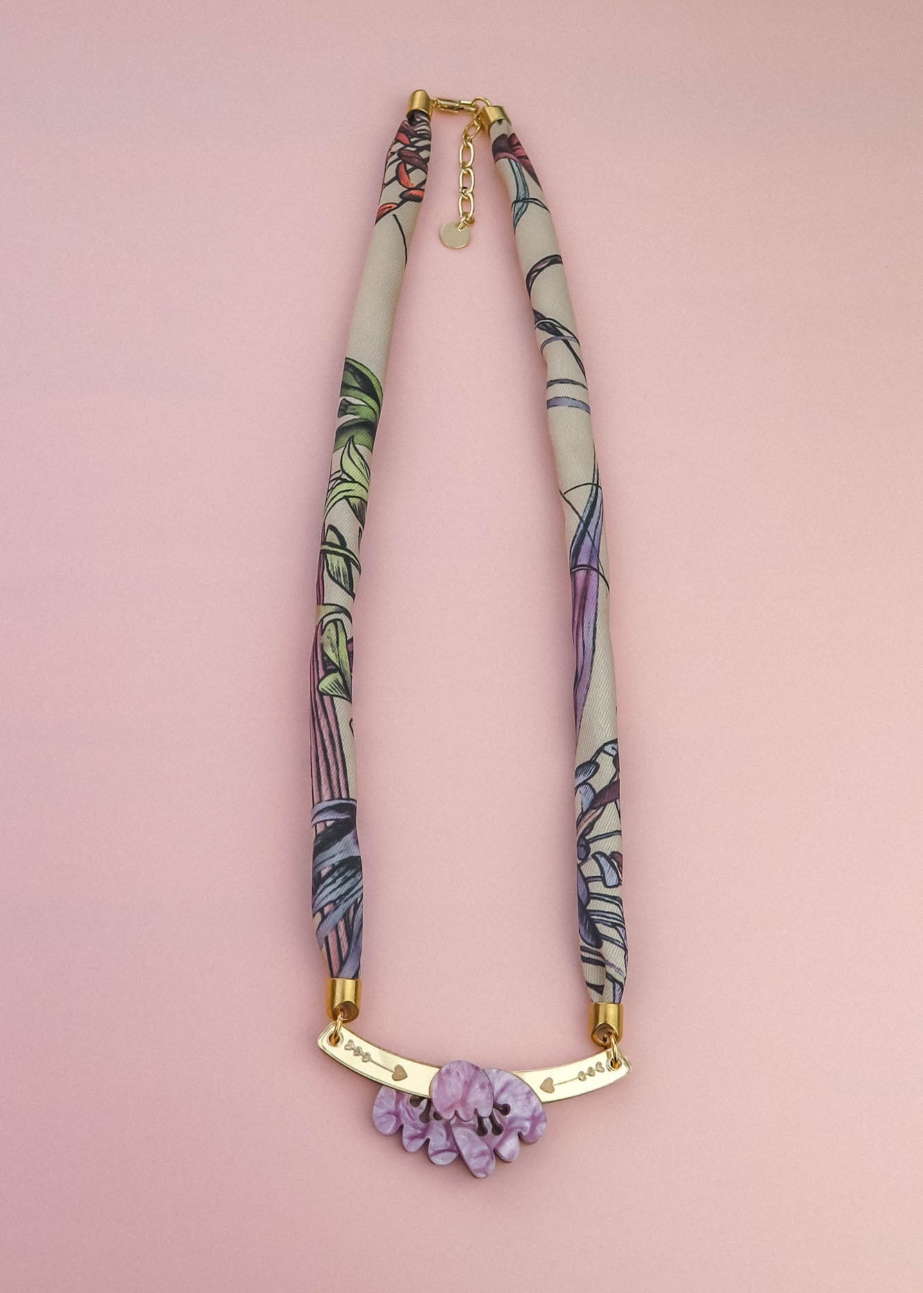 A handmade necklace with a fabric-wrapped band in muted colors and abstract patterns, featuring a gold bar centerpiece adorned with lilac stones, displayed on a light pink background—reminiscent of an elegant silk-print scarf.