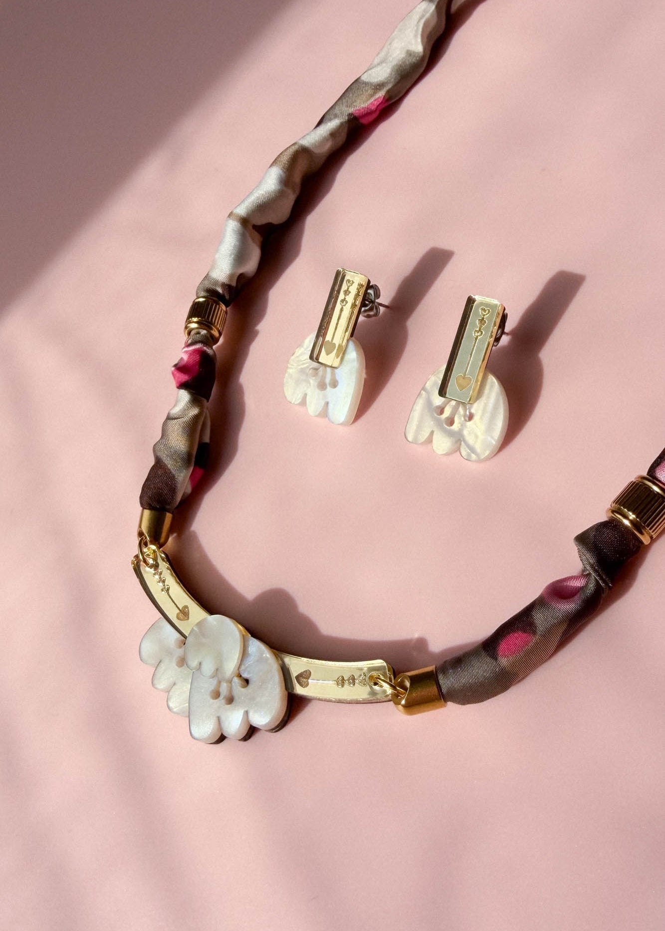 A handmade necklace and matching clip-on earrings with white, acrylic tulip-shaped pendants, gold accents, and a fabric-wrapped brown and pink chain, displayed on a soft pink background.