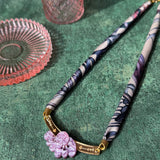 A handmade necklace featuring a patterned fabric and pink carved pendant with gold accents is displayed on a textured green surface, beside two pink glass dishes.