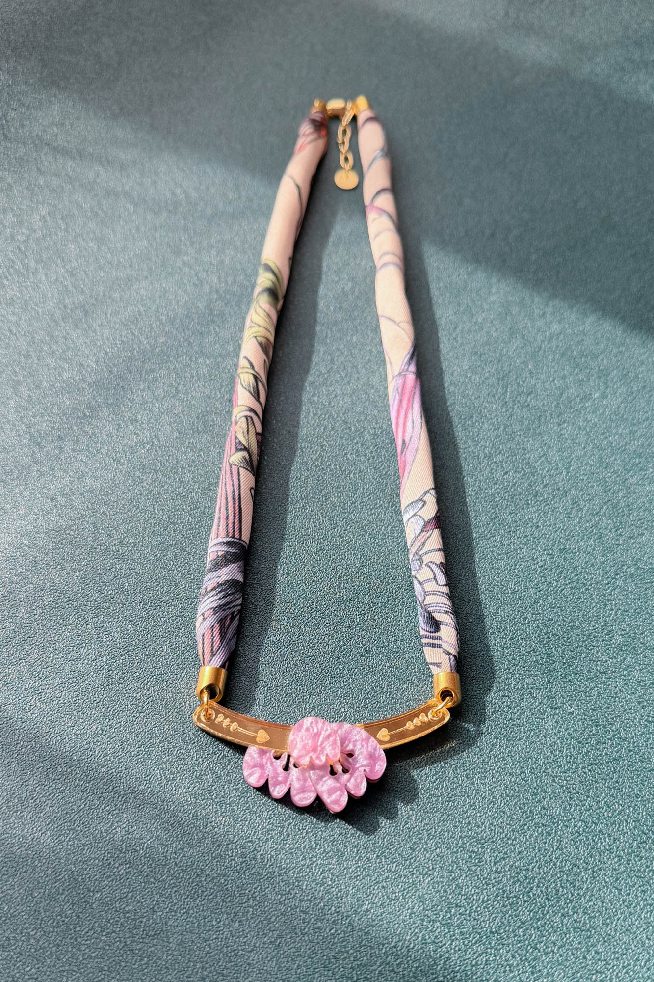 A handmade necklace with a fabric-wrapped band in pastel floral patterns, featuring a gold bar centerpiece adorned with pink flowers, and touches of acrylic tulip detail, displayed on a textured green surface.