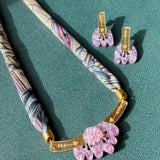 A handmade necklace with a patterned fabric cord, gold metal bar, and pink acrylic tulip pendants is displayed next to matching pink floral earrings on a textured teal surface.