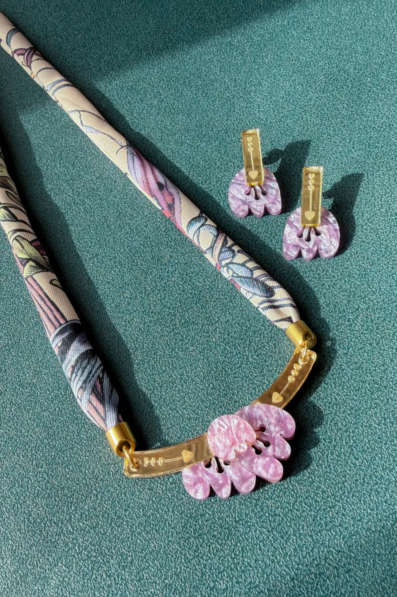 A handmade necklace with a patterned fabric cord, gold metal bar, and pink acrylic tulip pendants is displayed next to matching pink floral earrings on a textured teal surface.