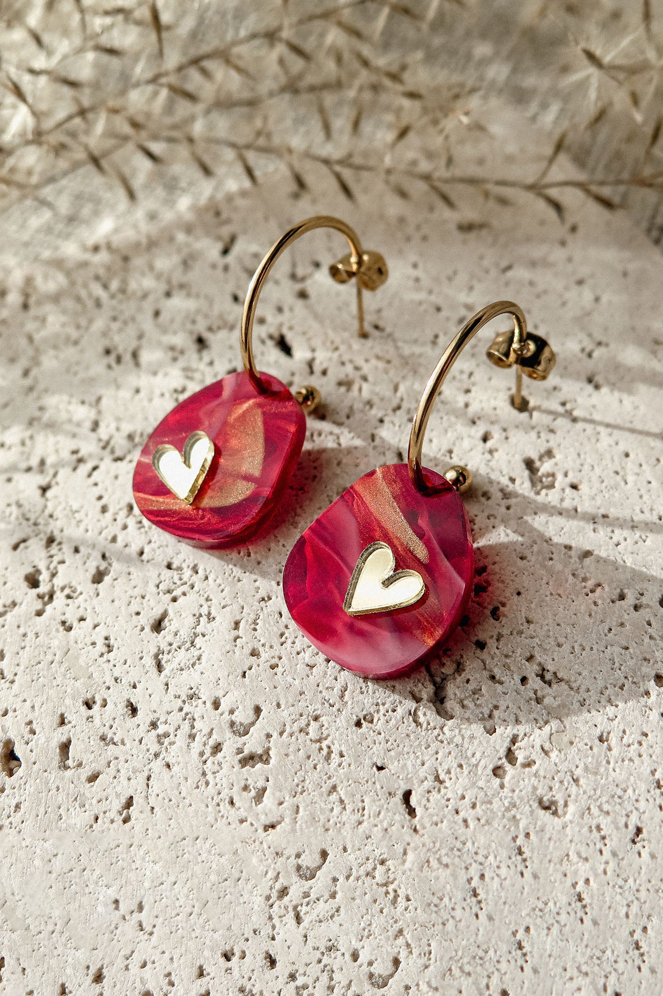A pair of gold hoop earrings with pink, marbled resin drops and small white heart charms, the Valentine Pebble Earrings are a romantic accessory displayed on a textured stone surface under natural light.