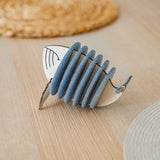 A decorative object resembling a stylized fish, made of alternating blue-grey felt and white pieces, sits on a light wooden table with woven placemats in the background, adding home charm and protecting surfaces like unique Whale Coasters.