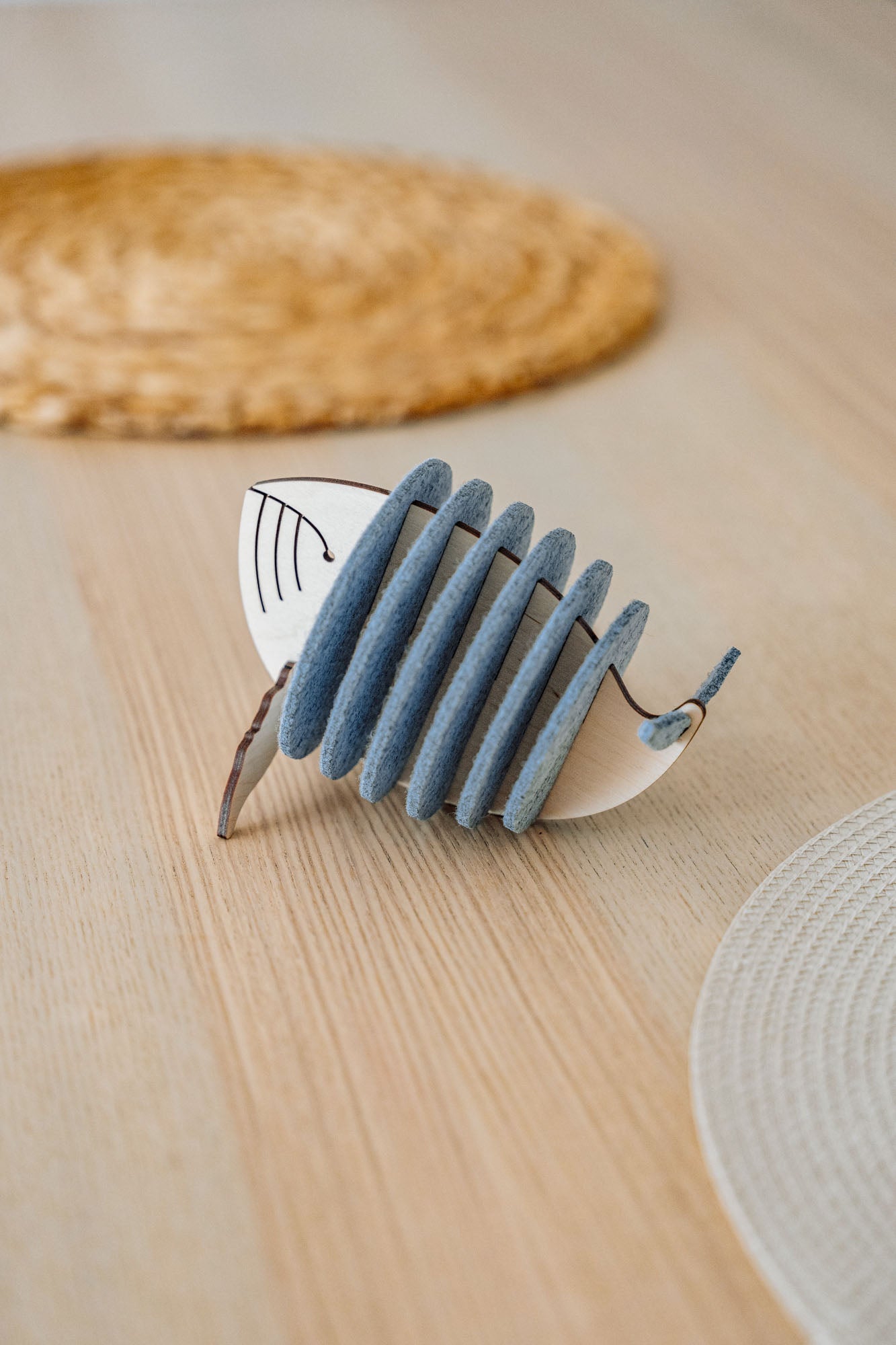 A decorative object resembling a stylized fish, made of alternating blue-grey felt and white pieces, sits on a light wooden table with woven placemats in the background, adding home charm and protecting surfaces like unique Whale Coasters.