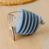 A wooden and felt napkin holder shaped like a fish sits on a light wooden table, adding home charm, with a woven placemat blurred in the background. Pair it with Whale Coasters to protect surfaces and complete your decor.