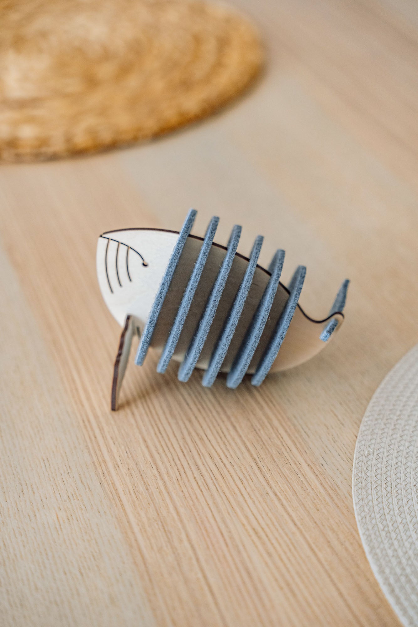 A wooden and felt Whale Coasters holder shaped like a stylized fish adds home charm as it sits on a light wooden table, protecting surfaces with woven placemats in the background.