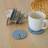 A white ribbed ceramic mug sits on a round gray felt coaster on a wooden table, with Whale Coasters and a whale-shaped holder nearby. A woven placemat adds home charm while these coasters help protect surfaces in style.