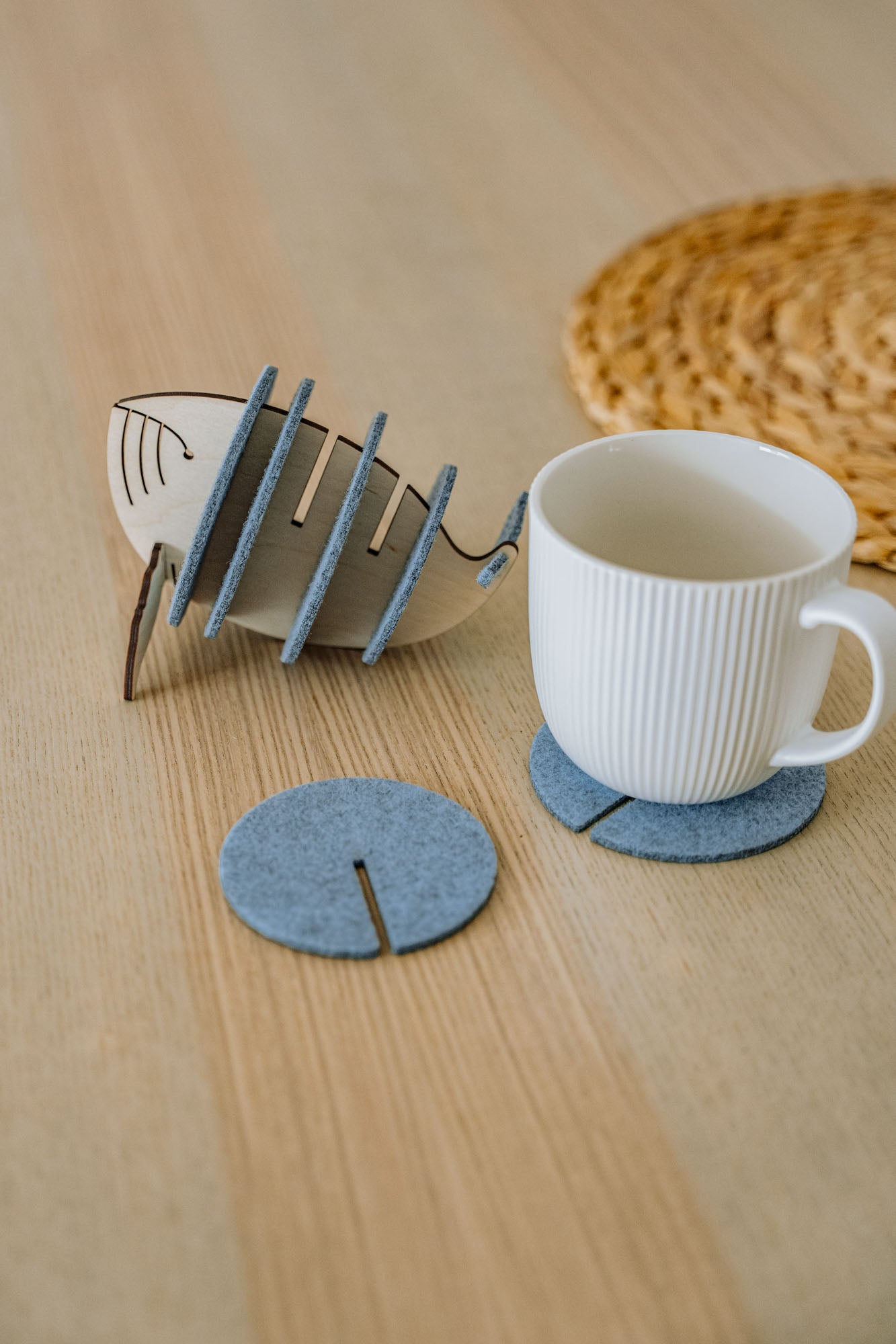 A white ribbed ceramic mug sits on a round gray felt coaster on a wooden table, with Whale Coasters and a whale-shaped holder nearby. A woven placemat adds home charm while these coasters help protect surfaces in style.