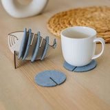 A white ribbed mug sits on a round gray felt coaster, designed to protect surfaces. Nearby, more Whale Coasters are stacked in a fish-shaped holder, with woven and ceramic decor adding home charm in the background.